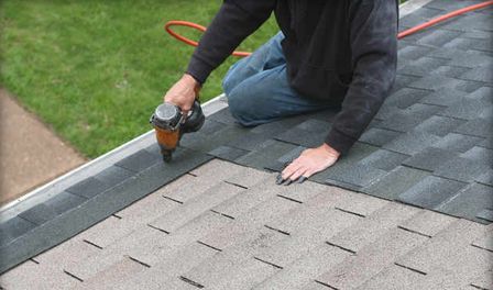 Person kneeling on a roof, using a nail gun to install shingles.