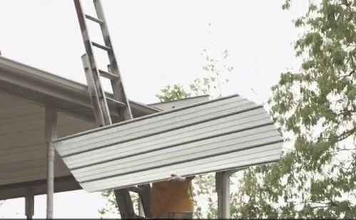 Person on ladder holding a large, white panel near a building's roof with trees in the background.
