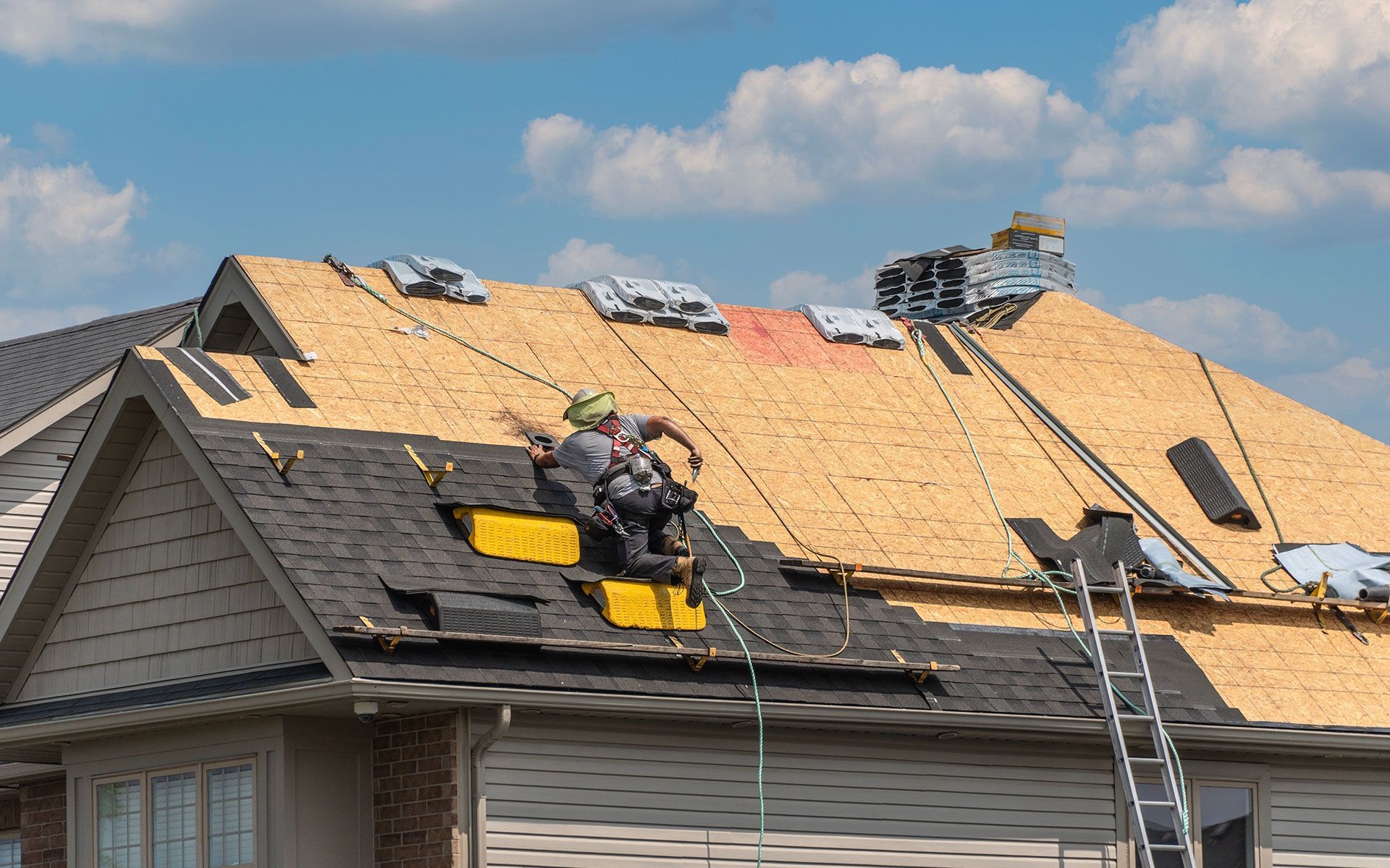 Roofers working on a house roof. One is secured by a safety harness. Blue sky in background.