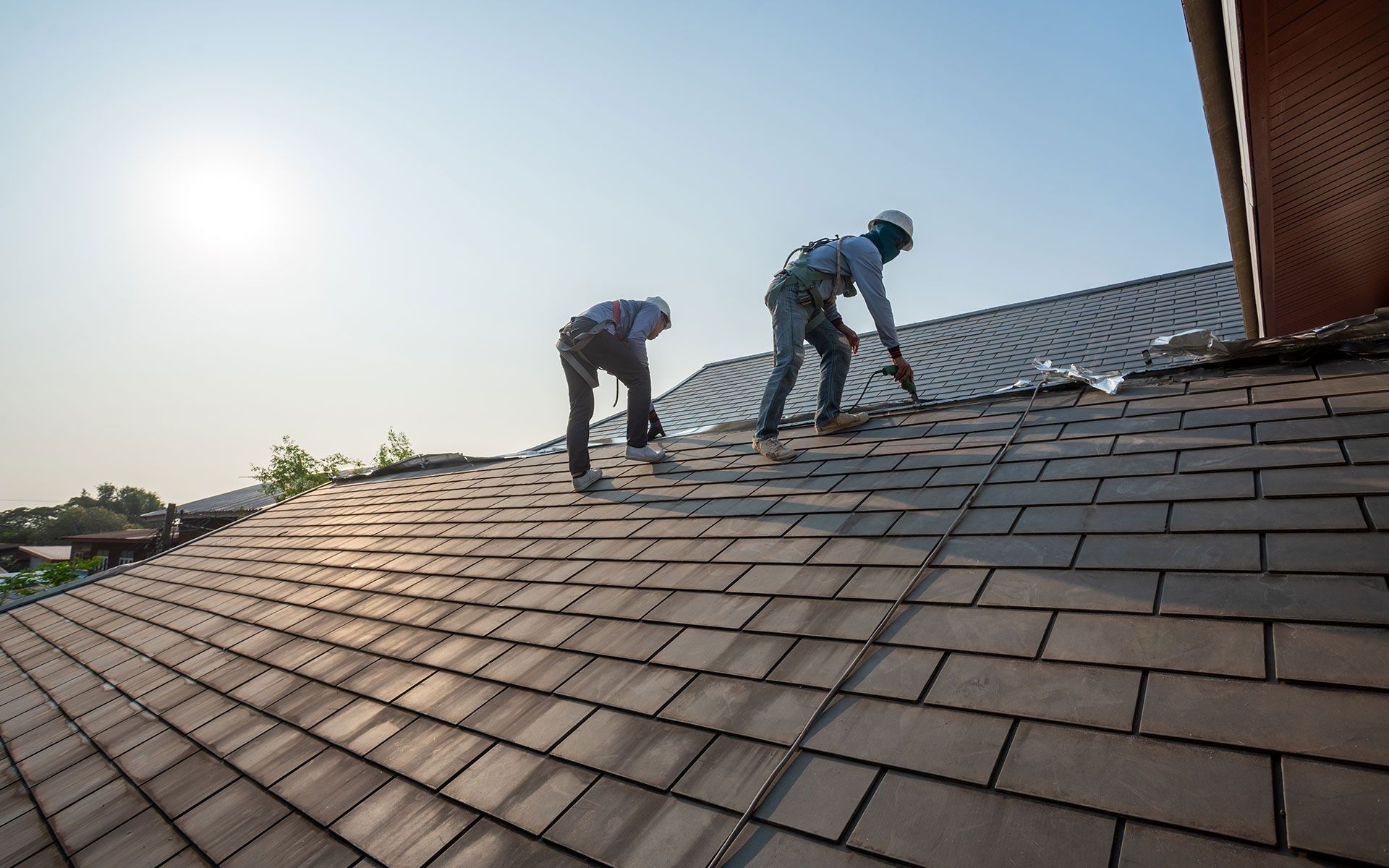 Two roofers working on a shingled roof under a bright sky.