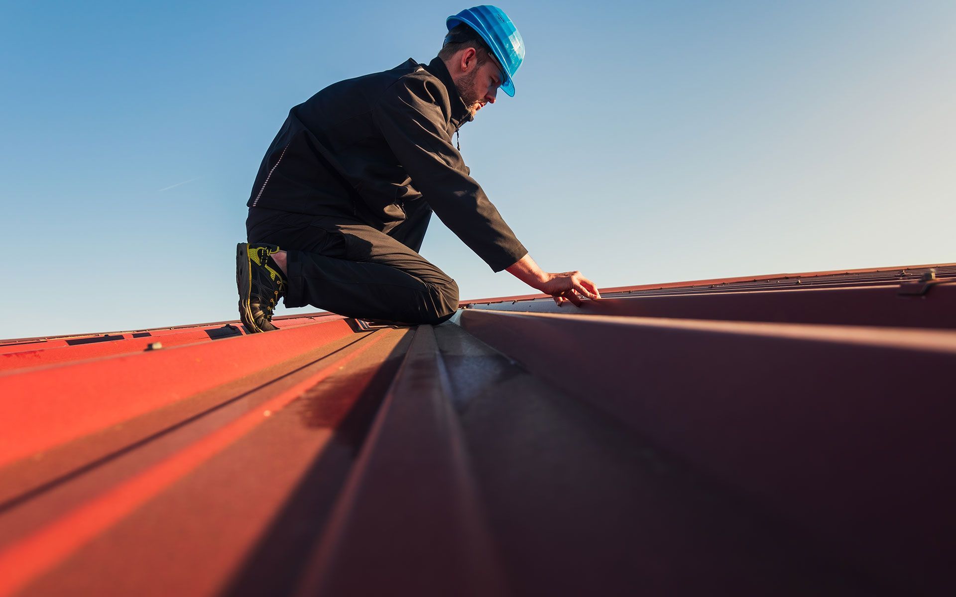 Roofer in blue hard hat on red metal roof, inspecting the surface under a clear blue sky.