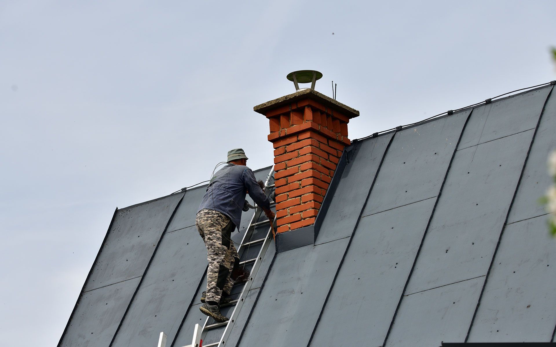 Person on a ladder, ascending to a brick chimney on a gray metal roof.
