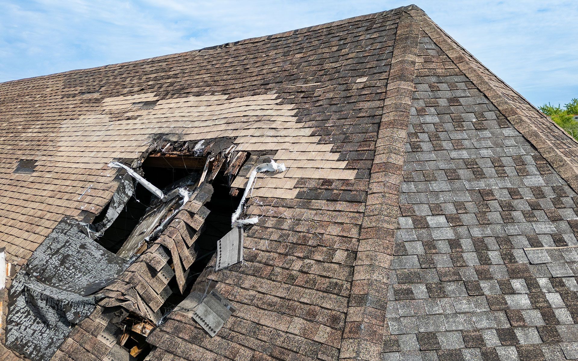 Damaged asphalt shingle roof with a large hole and missing sections.