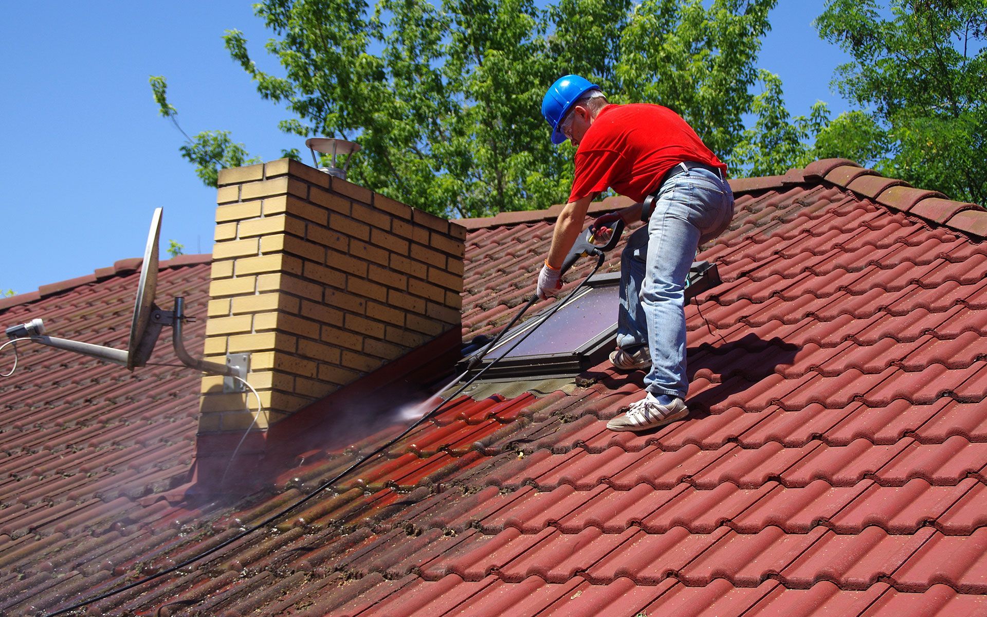 Man pressure washing red tiled roof, near chimney and satellite dish. Bright sun, clear blue sky.