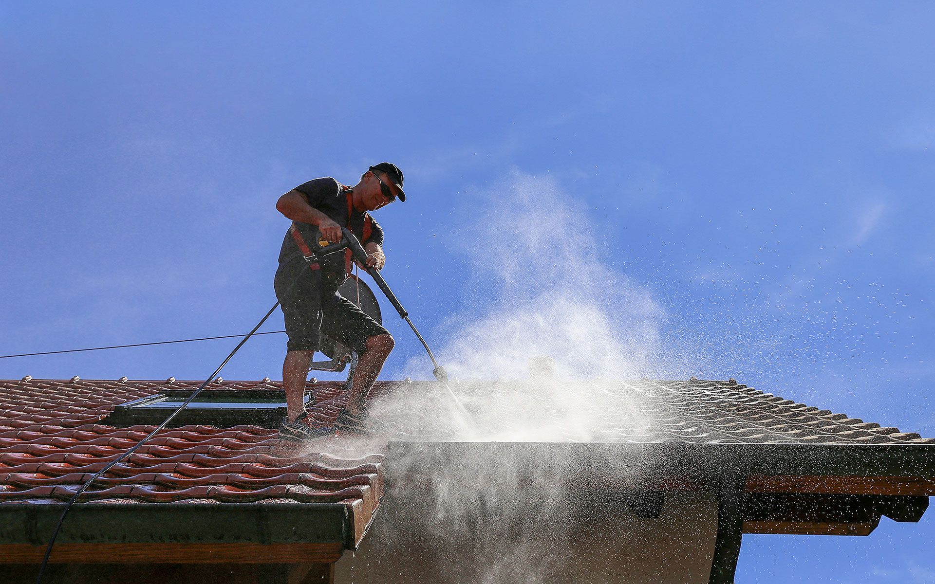 Person power washing a tiled roof on a sunny day, spraying water.