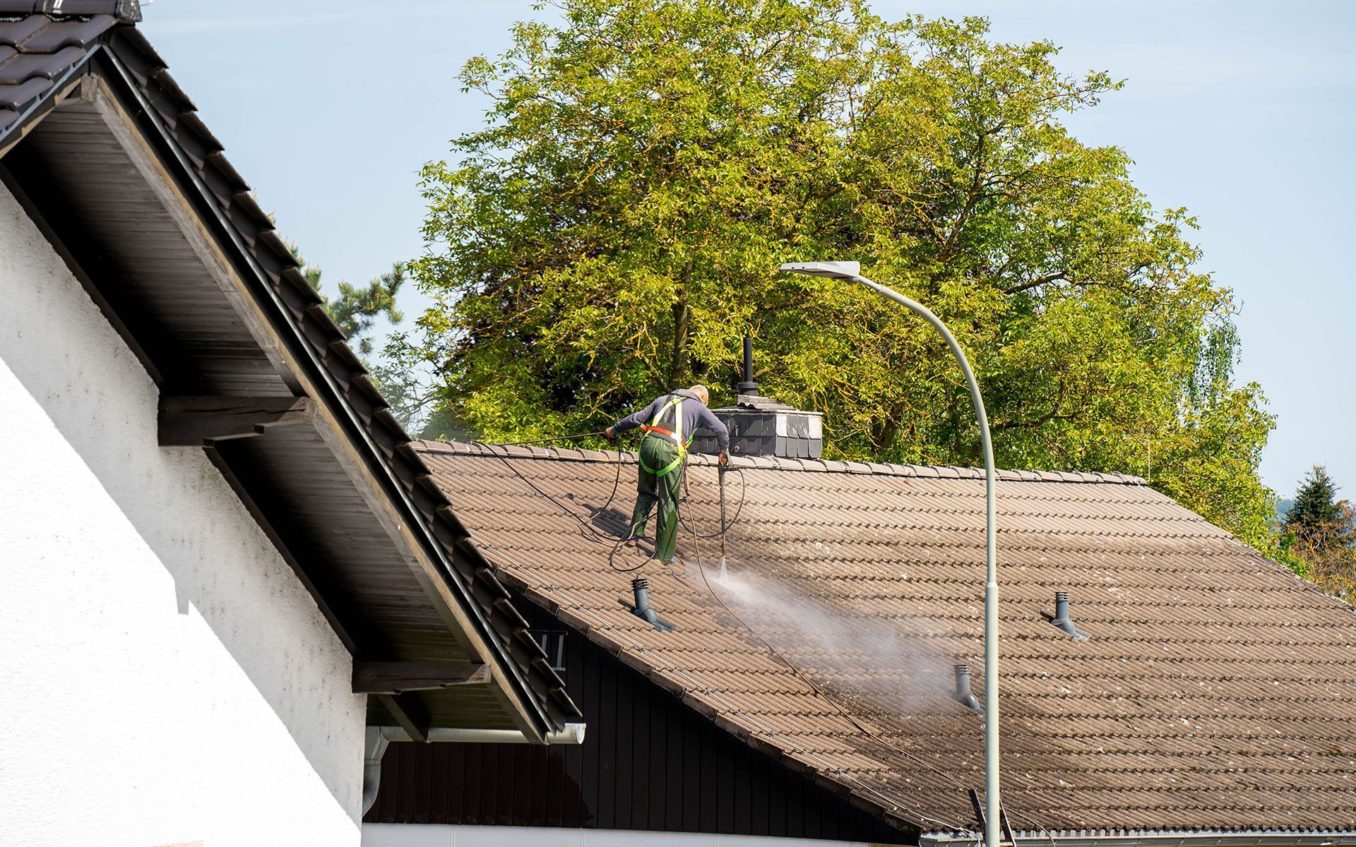 Person pressure washing a brown shingled roof, trees in the background.