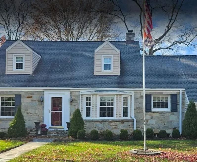 Stone house with dormers, shutters, and American flag on a grassy lawn.