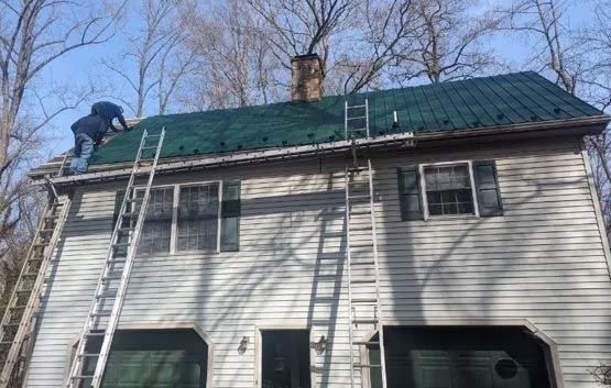 Person on a roof with green metal roofing; ladders against the house.