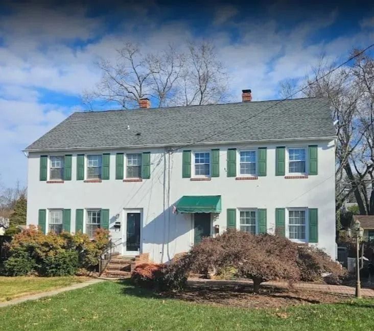 White two-story building with green shutters and awning. Green lawn and leafless tree in front. Blue sky.