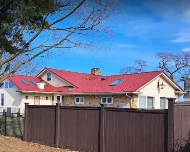 House with a red metal roof behind a brown fence, under a blue sky.