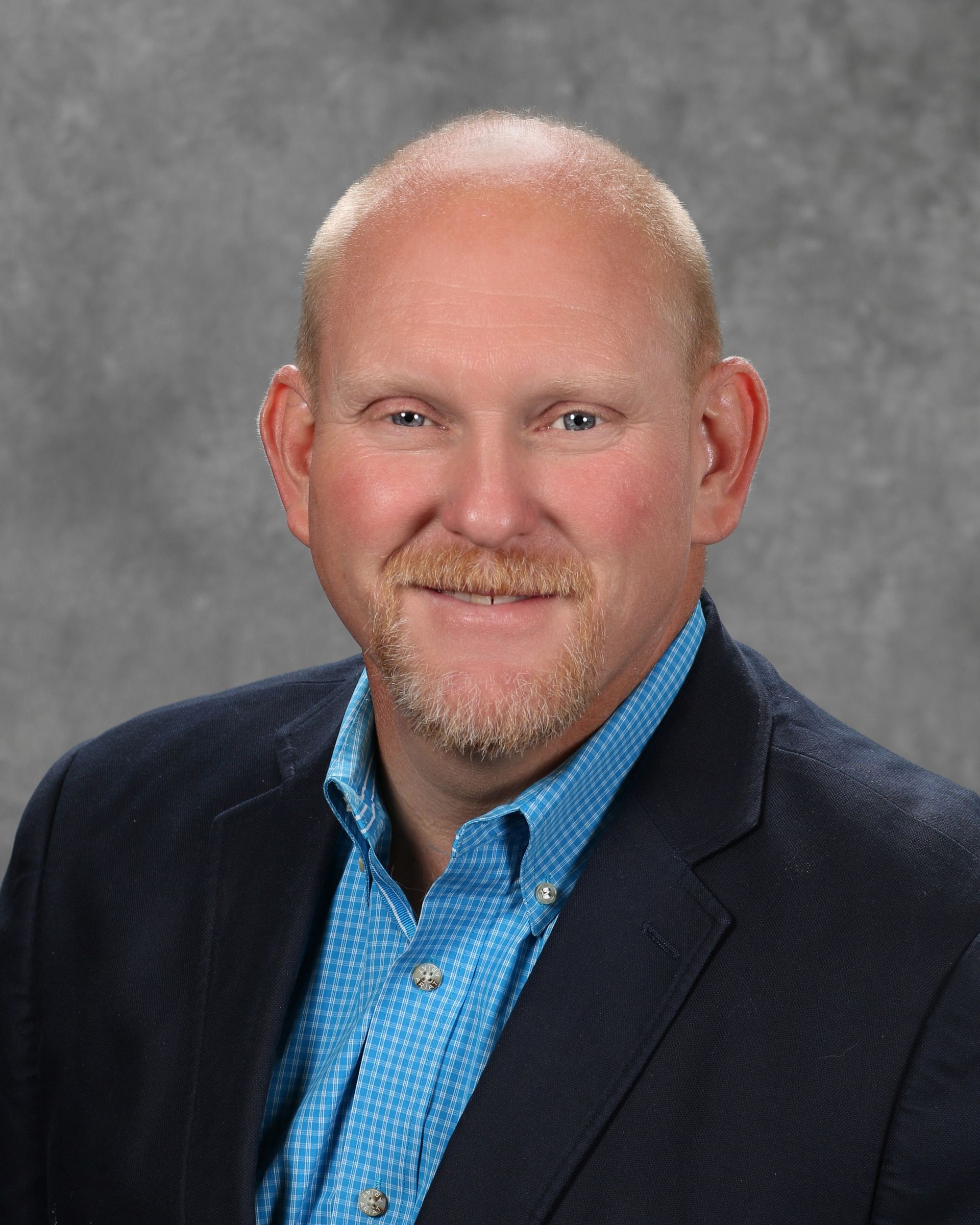 Man with fair skin, smiling, wearing a blue button-down shirt and navy blazer; gray background.