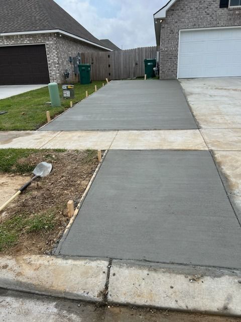 Newly poured concrete driveway and sidewalk next to a house. A shovel rests in the grass.