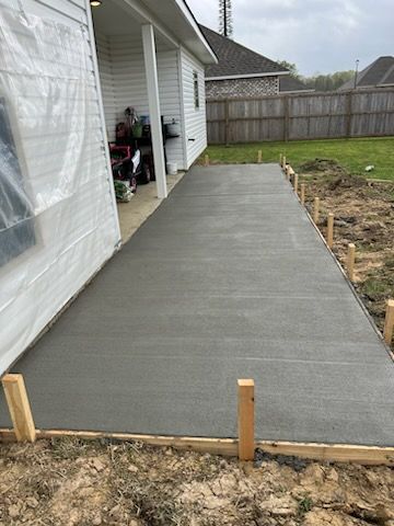 Newly poured concrete patio beside a white house, framed with wooden boards in an outdoor setting.