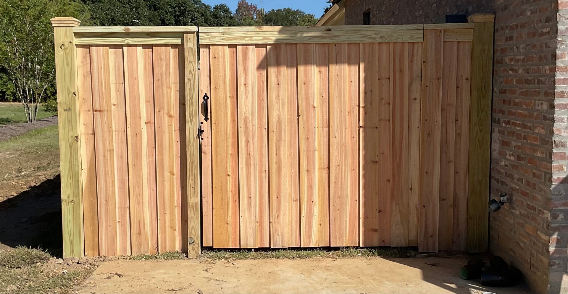 Wooden gate and fence, light brown wood, green posts, set against a brick wall and grassy area.