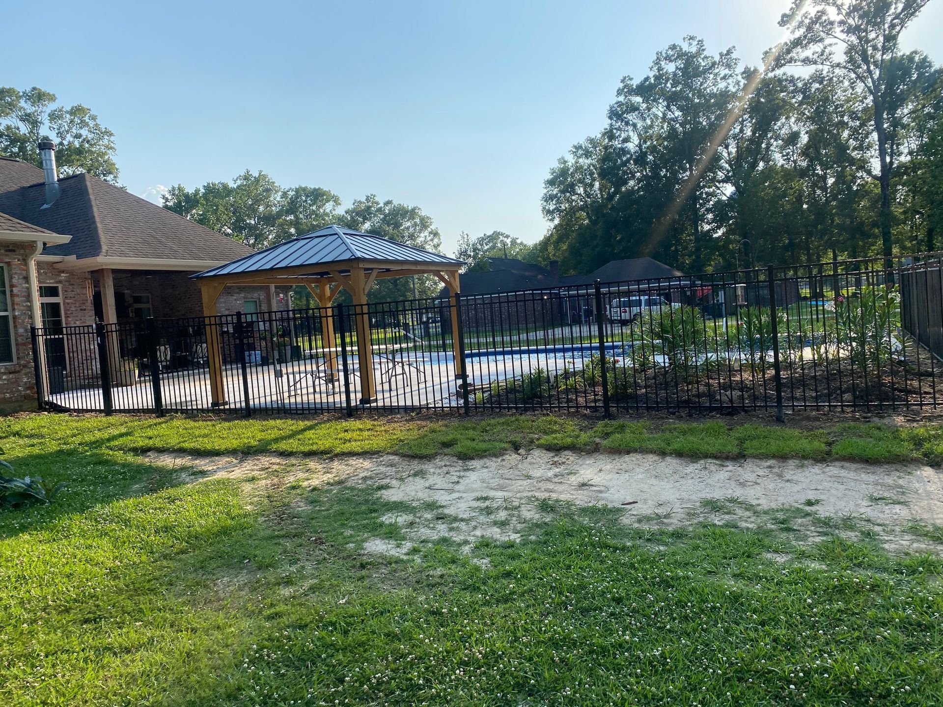 A fenced backyard with a gazebo by a pool, viewed from the grass, under a bright sky.