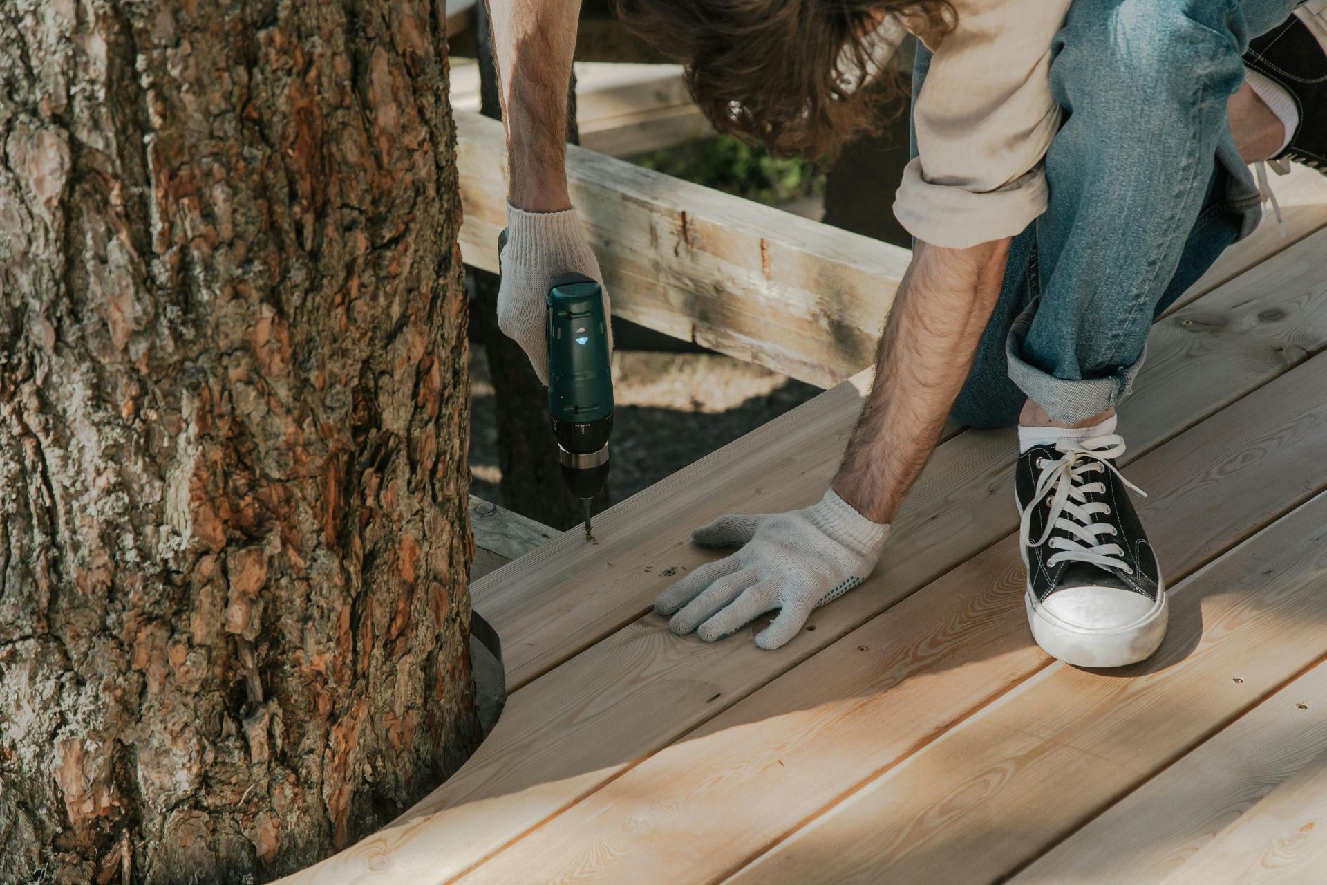 Person using a drill to attach wooden planks for a deck. They are wearing gloves, jeans, and sneakers next to a tree.