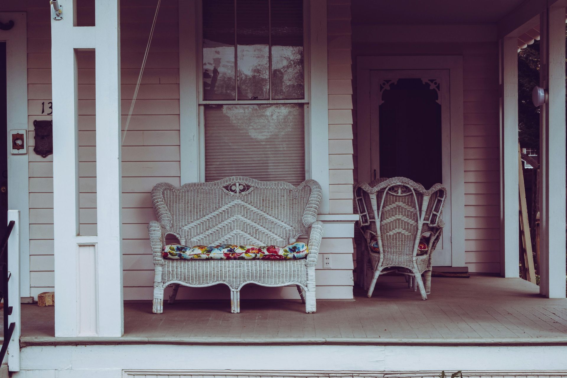 Wicker furniture on a porch, white exterior. Bench with floral cushion, armchair. Window, door.