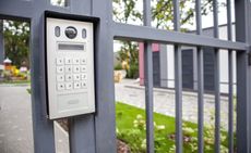 Security keypad and camera on a metal gate, in front of a modern building.