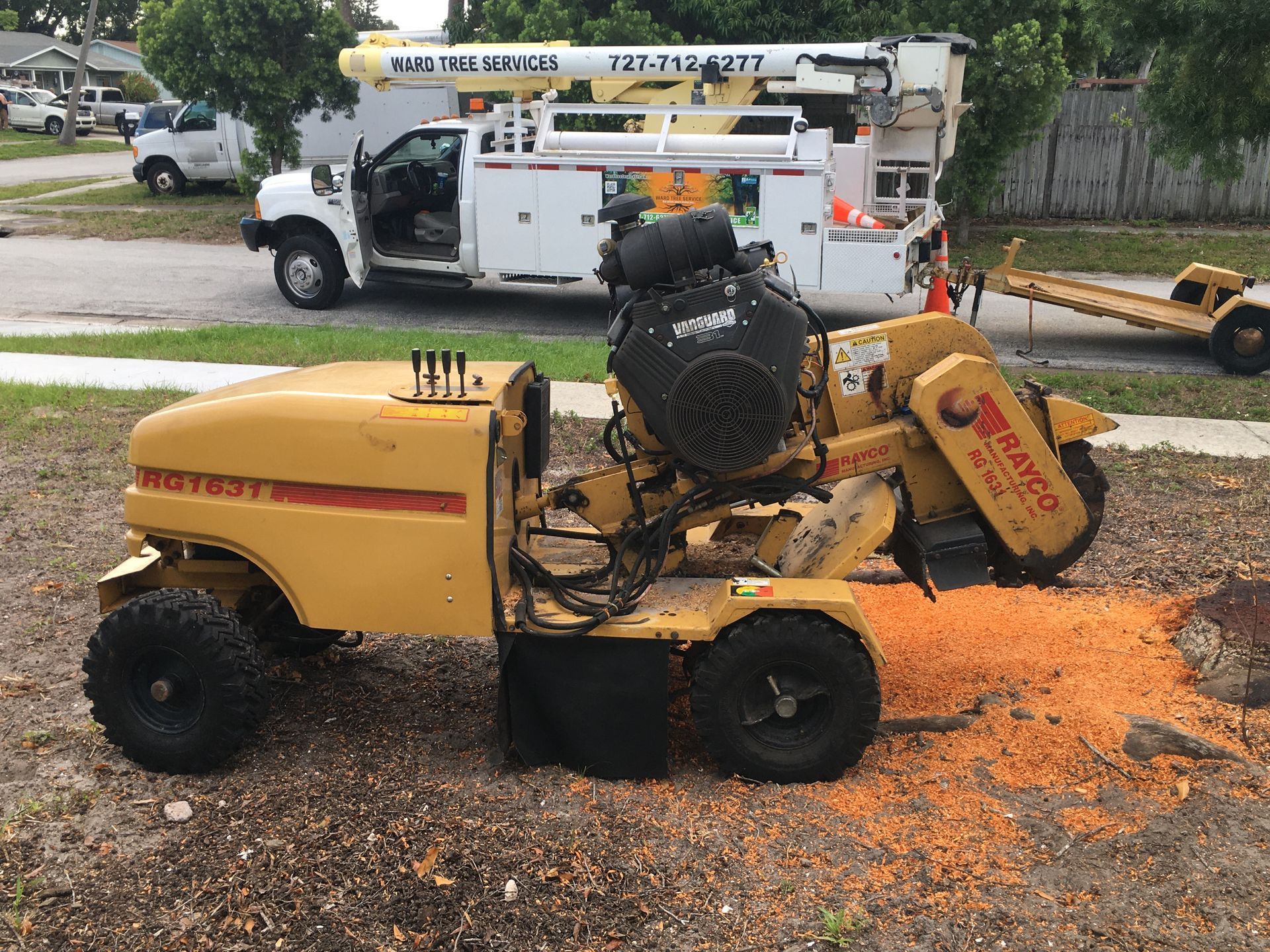 An excavator bucket digs into the soil, removing tree stumps and roots from a grassy yard.