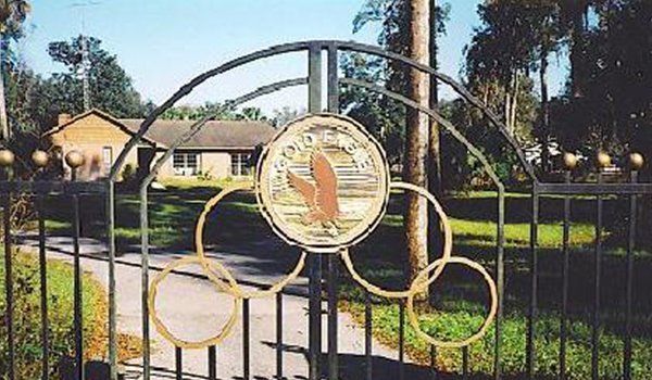 Black metal gate with a circular seal, opening to a green lawn and a house in the background