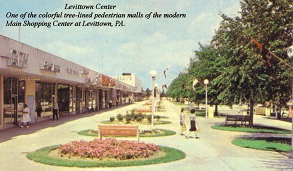 Historic Levittown Center shopping plaza with tree-lined walkway, storefronts, benches, and flower beds