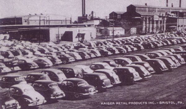Rows of vintage cars parked in a factory lot beside industrial buildings.
