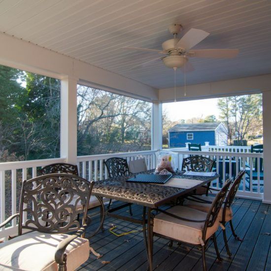 Covered porch with metal table and chairs, ceiling fan, and a blue house visible beyond the railing.