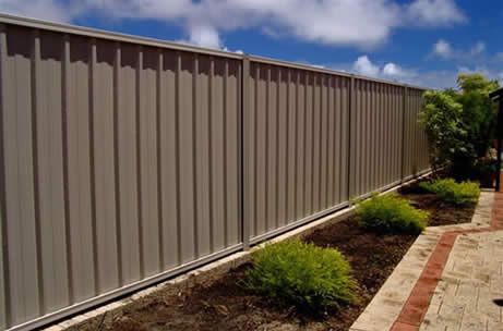 A tall beige corrugated metal privacy fence lines a garden bed with small shrubs, next to a paved brick path under a blue sky.