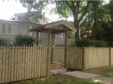 A wooden picket fence and a covered entry gate leading to a light-colored house with a porch.