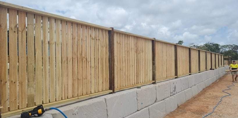 A wooden fence sits atop a long concrete block retaining wall outdoors under a cloudy sky.