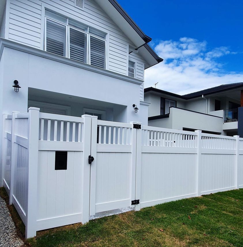 A white vinyl privacy fence with a gate stands in front of a modern white two-story house under a blue sky.