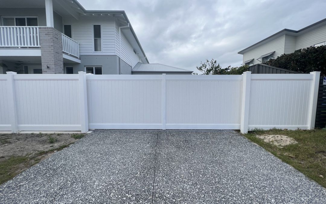 A white, solid privacy fence with double gates installed across a grey gravel driveway in front of a house.