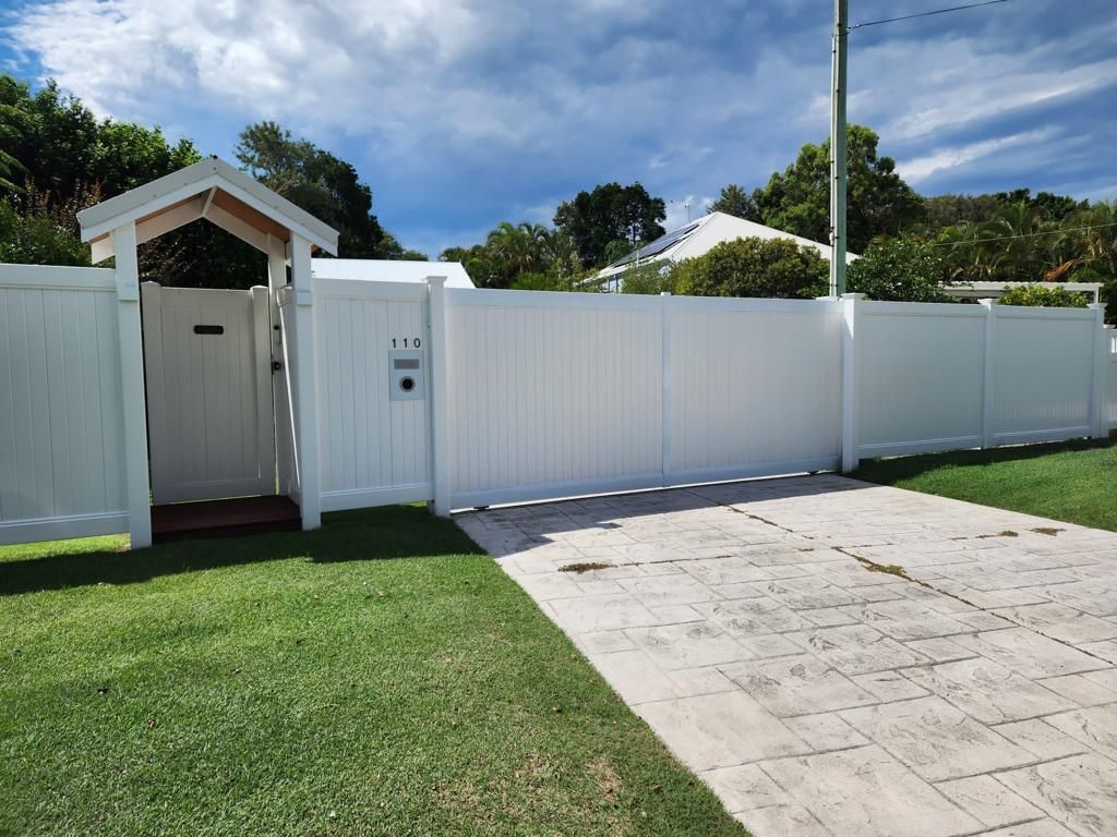 A white vinyl fence with a matching pedestrian gate and driveway entrance, situated along a paved driveway and lawn.
