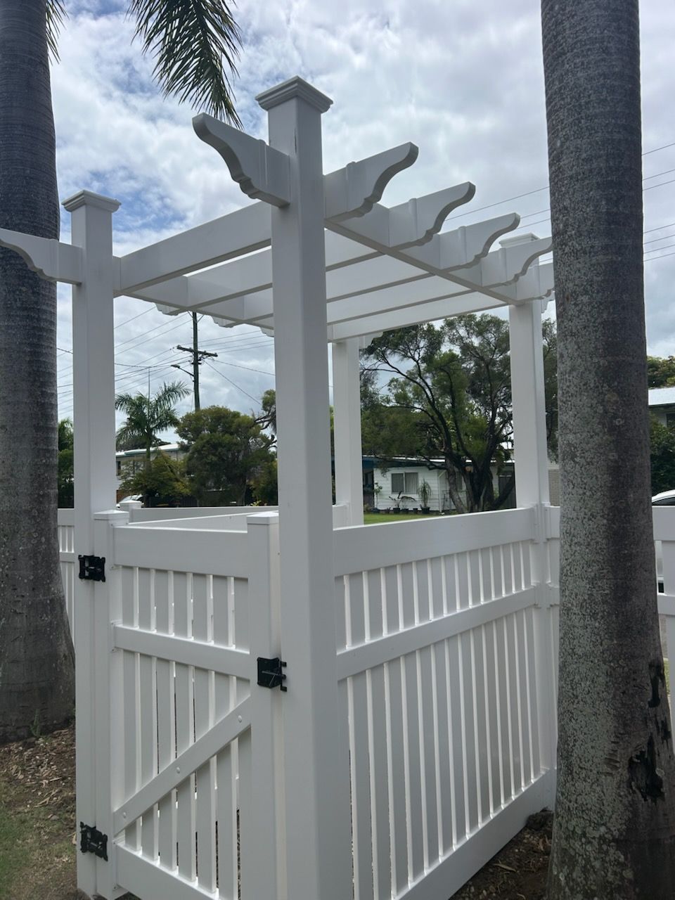 White wooden pergola gate with a fence, set between two palm trees.