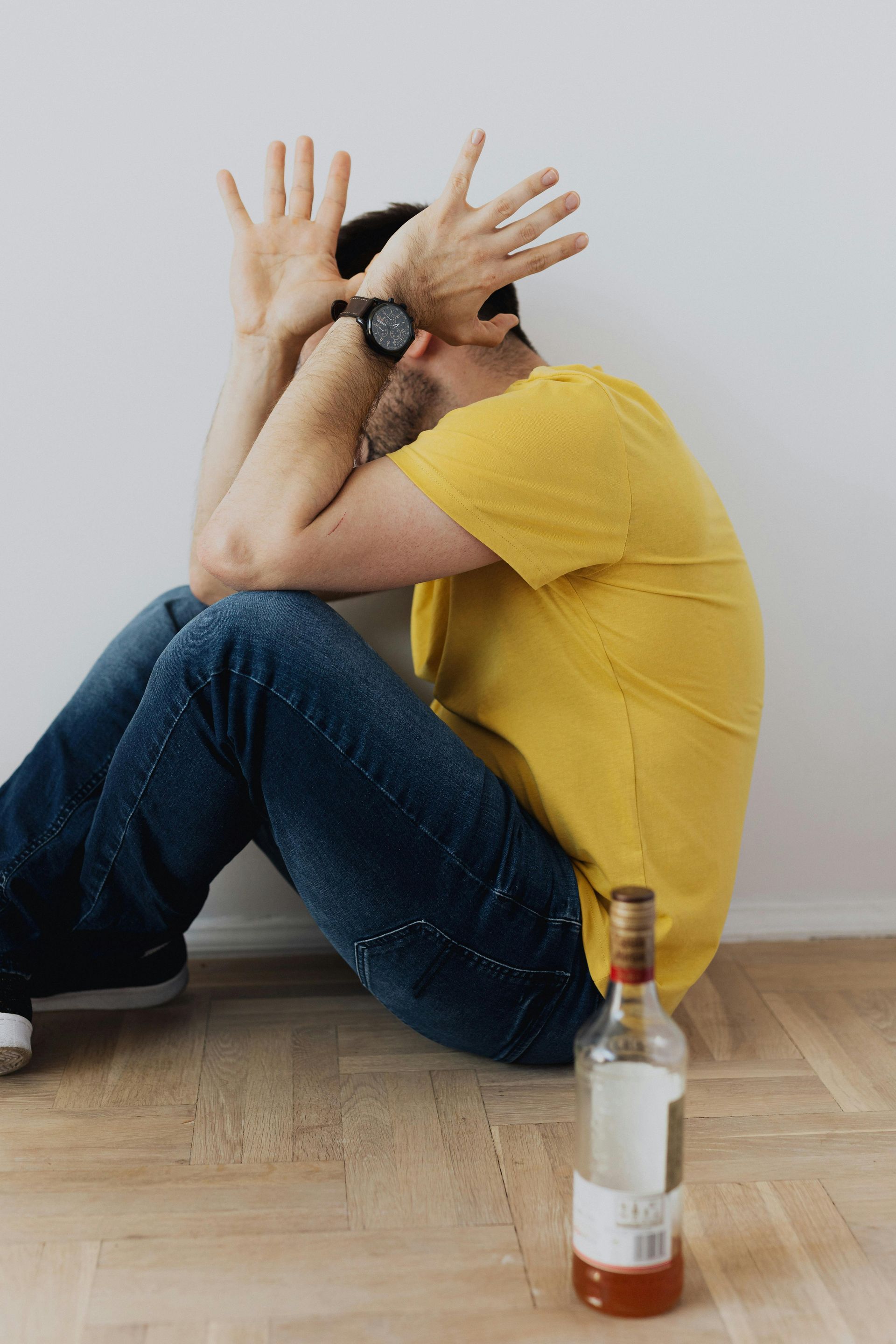 Man sitting on the floor with hands over his face, bottle of alcohol nearby, against white wall.