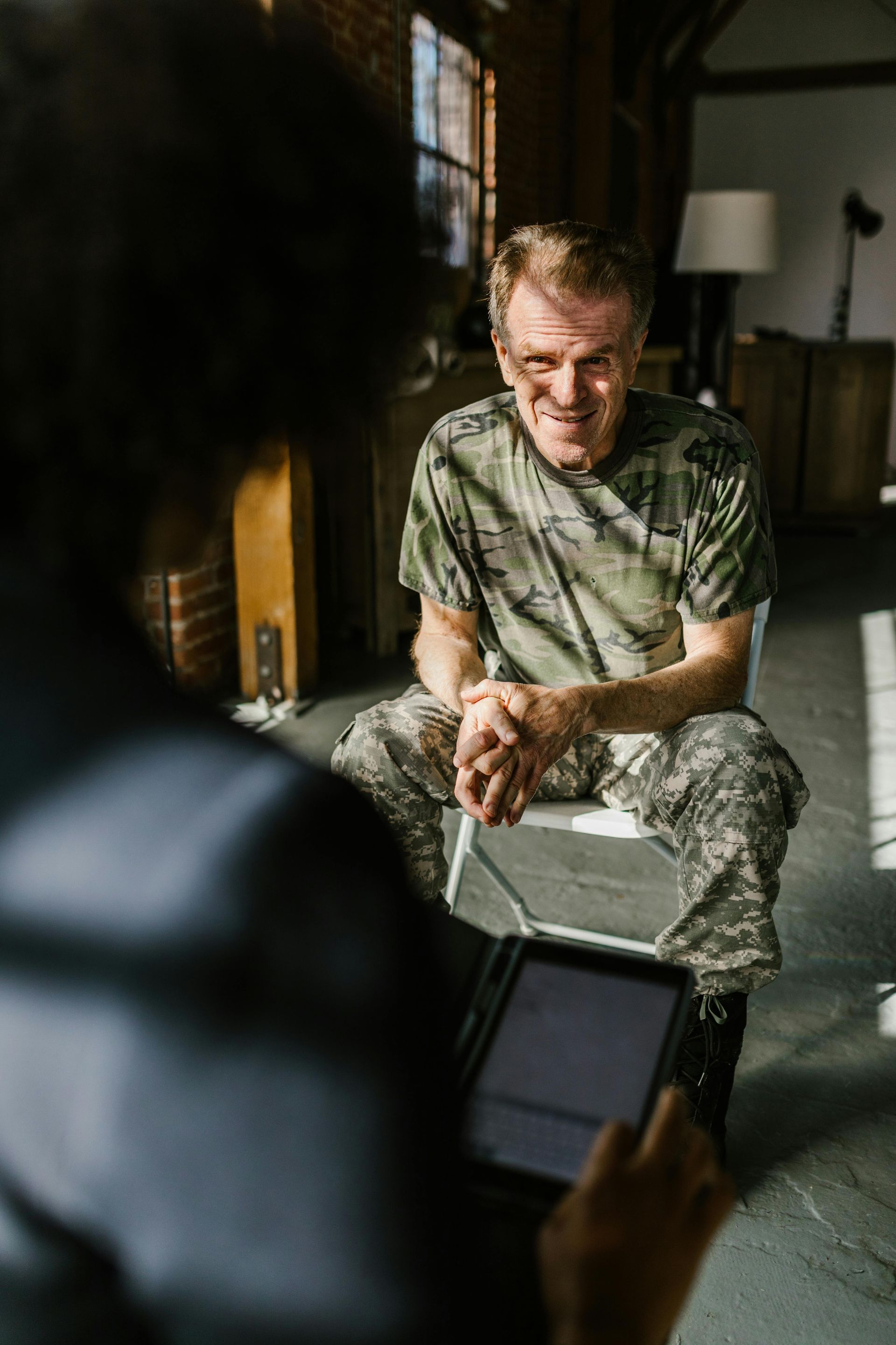 Man in camouflage shirt seated, facing person holding a tablet. Indoor setting, sunlight.