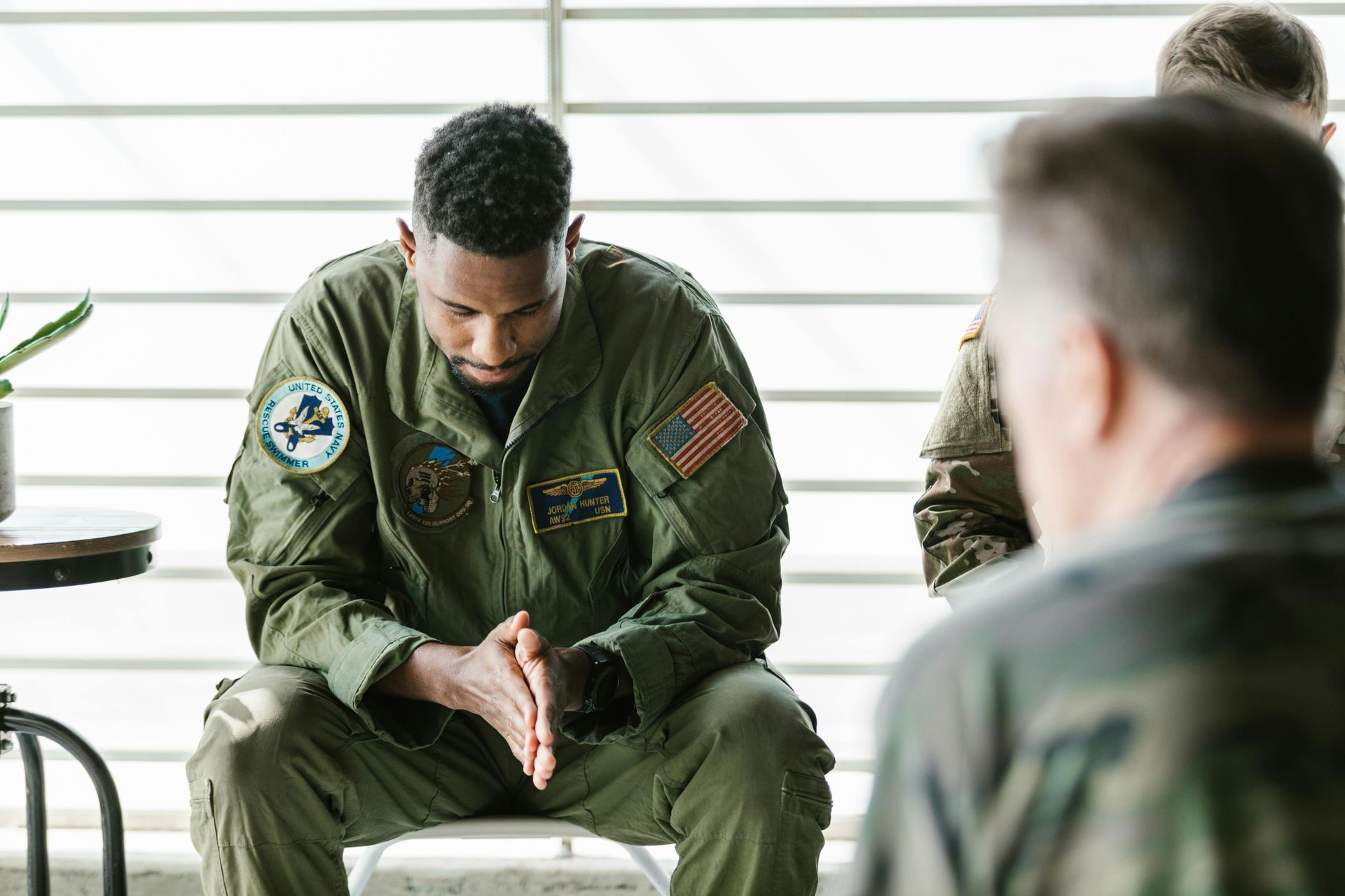Man in military uniform, head bowed, clasped hands, seated in a group setting.