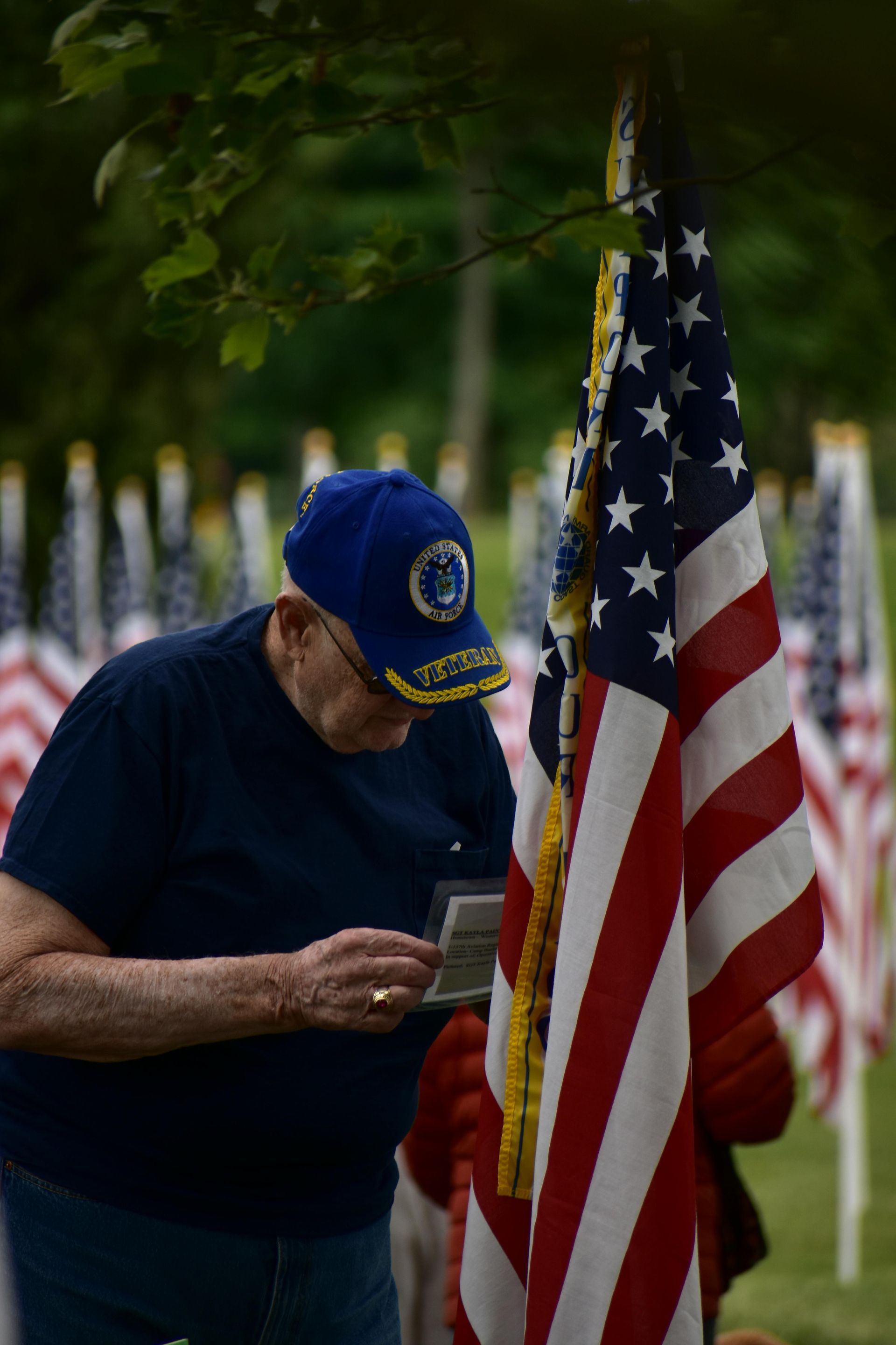 Man in blue cap examines a card near a U.S. flag at a memorial. Many flags in background.