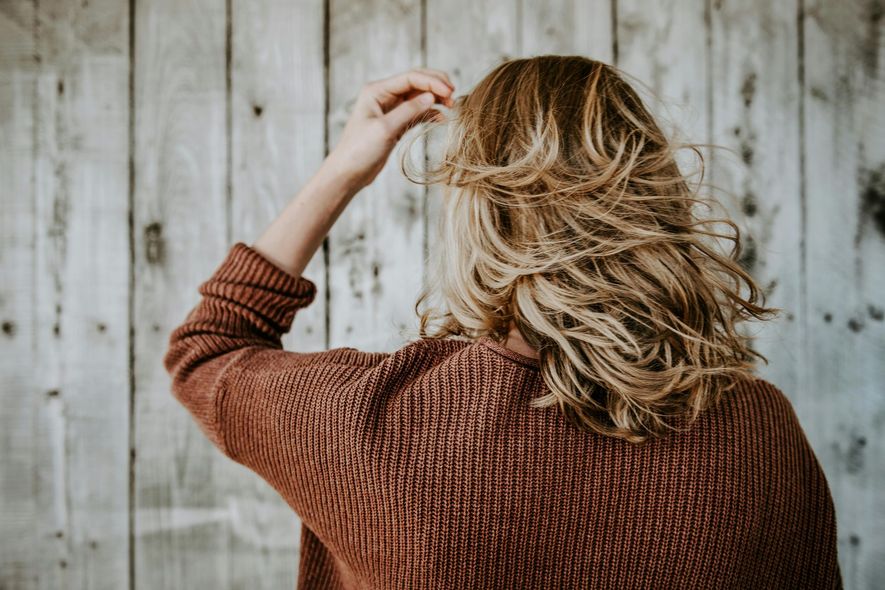 A woman in a brown sweater is touching her hair in front of a wooden wall.