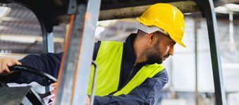 A man wearing a hard hat and safety vest is driving a forklift in a factory.