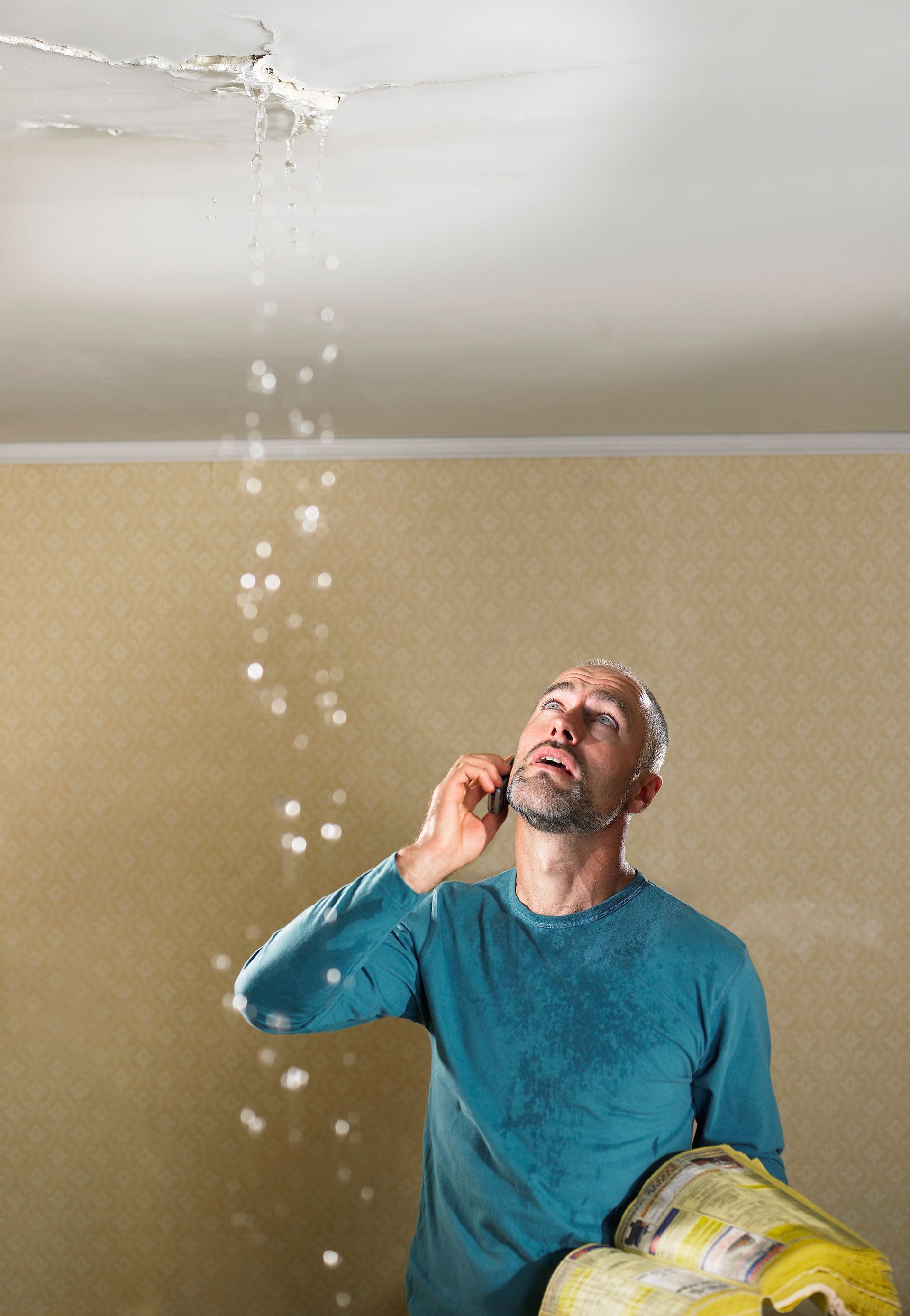 Man in blue shirt on phone looking up at water leaking from the ceiling.