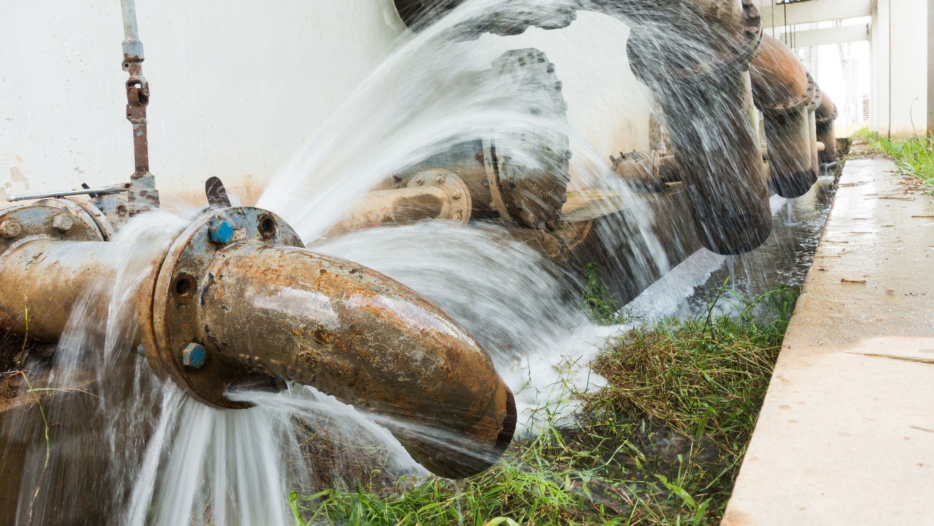 Rusted pipes burst, spewing water, in a grassy area next to a building.