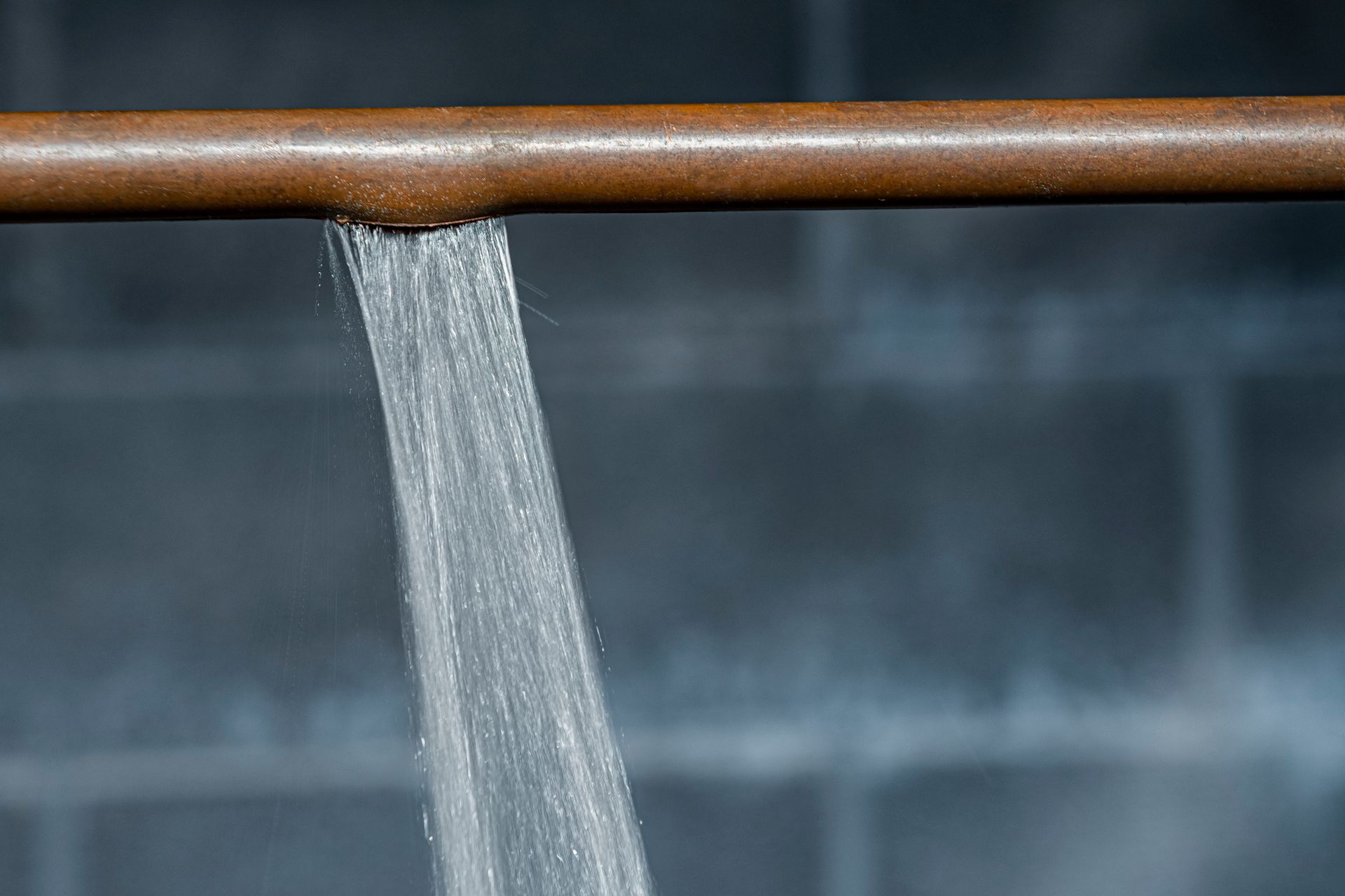 Water gushing from a split copper pipe, set against a gray brick wall.