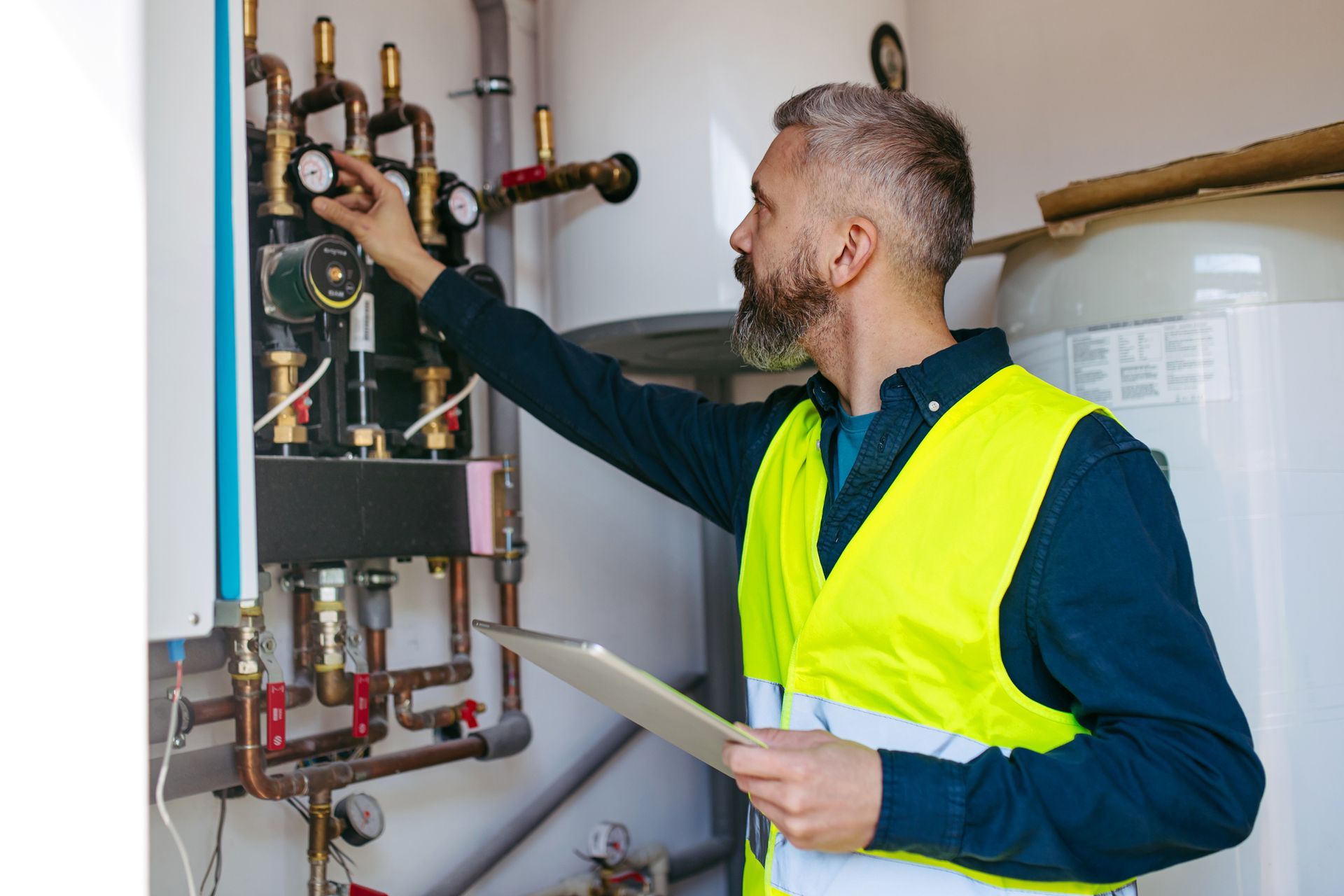 Man in a safety vest inspects plumbing with a tablet in a boiler room.