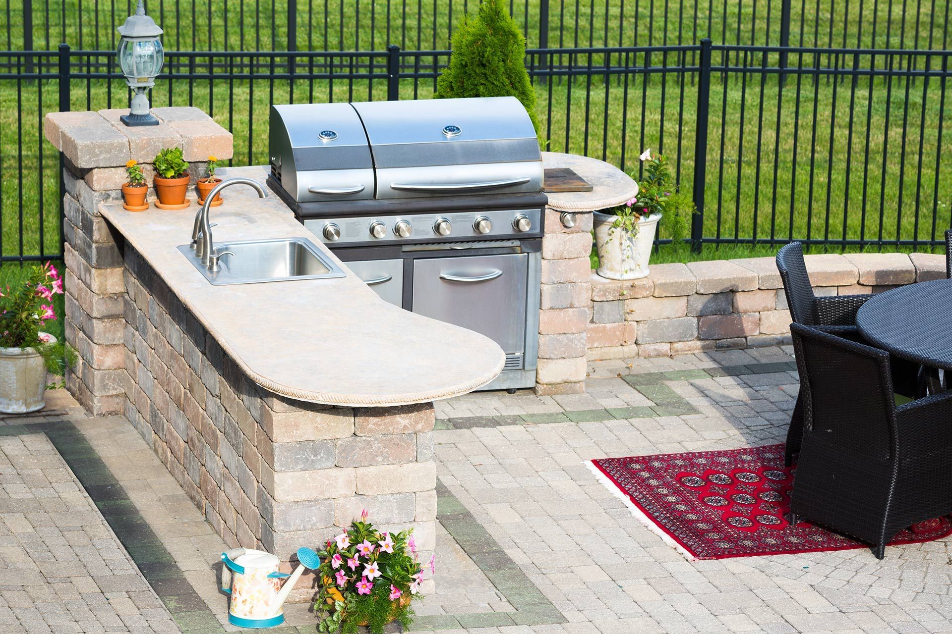 An outdoor kitchen with a stainless steel grill, stone counter, sink, and a paved patio area with a table and rug.