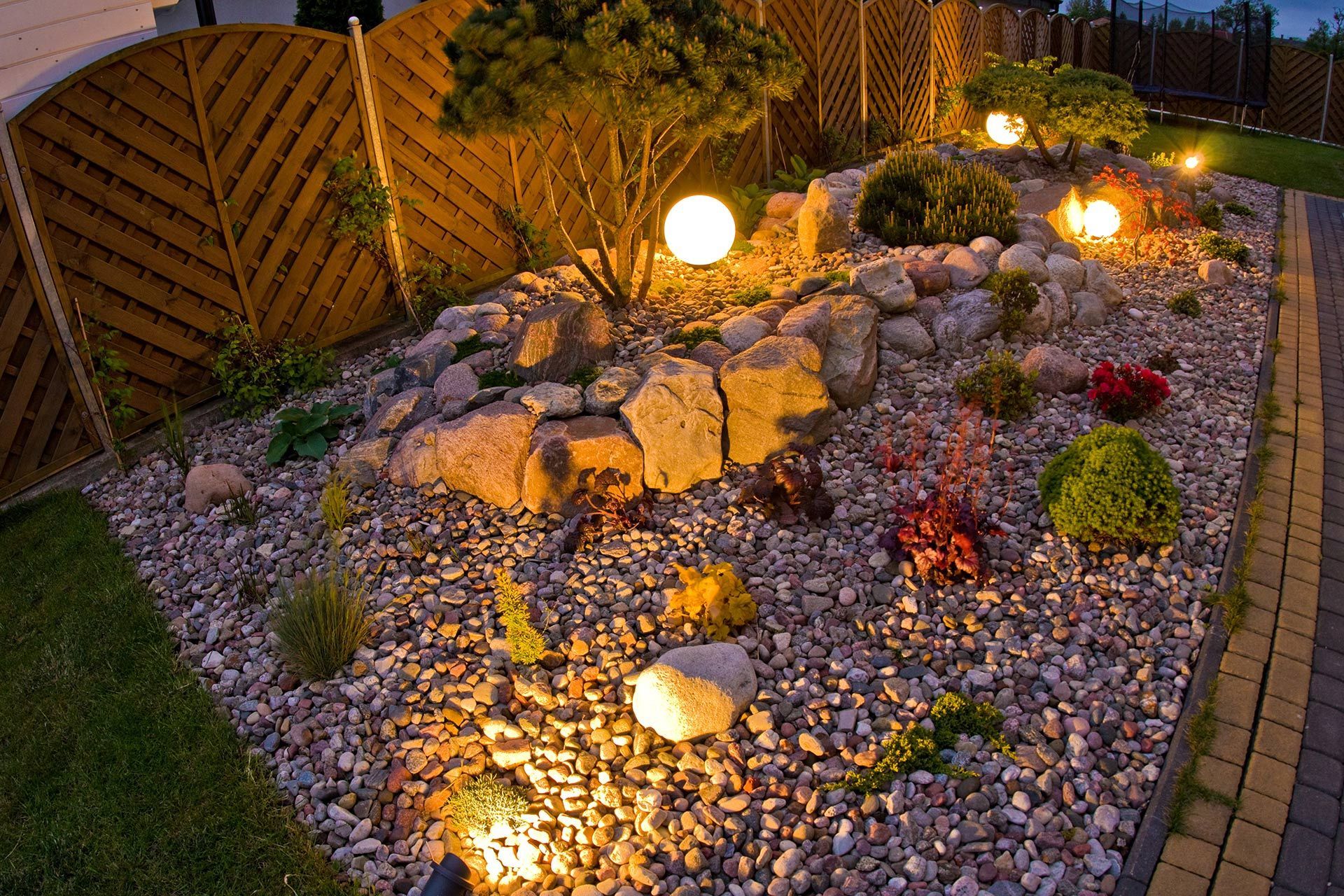 A landscaped garden at dusk featuring glowing spherical lights, rocks, small shrubs, and a decorative wooden fence.