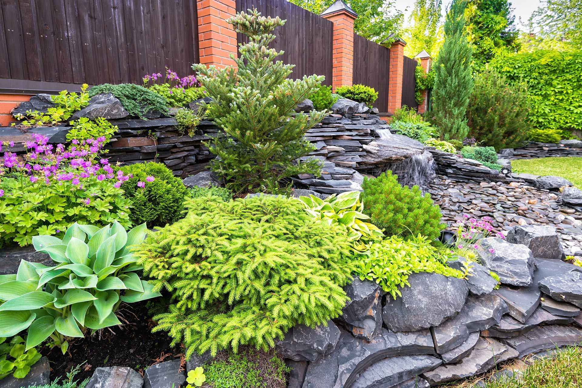 A tiered rock garden features lush green shrubs, purple flowers, and a coniferous tree against a brick and wood fence.