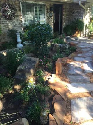 A flagstone path leads to a house entrance with a brick exterior, stone landscaping, and various shrubs and plants.