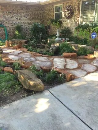 A flagstone pathway leads to a garden bed with rocks, shrubs, and a bird bath against a brick house.