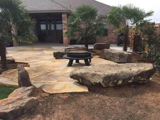 A backyard patio featuring a stone floor, a small round table, and large accent boulders near a brick house.